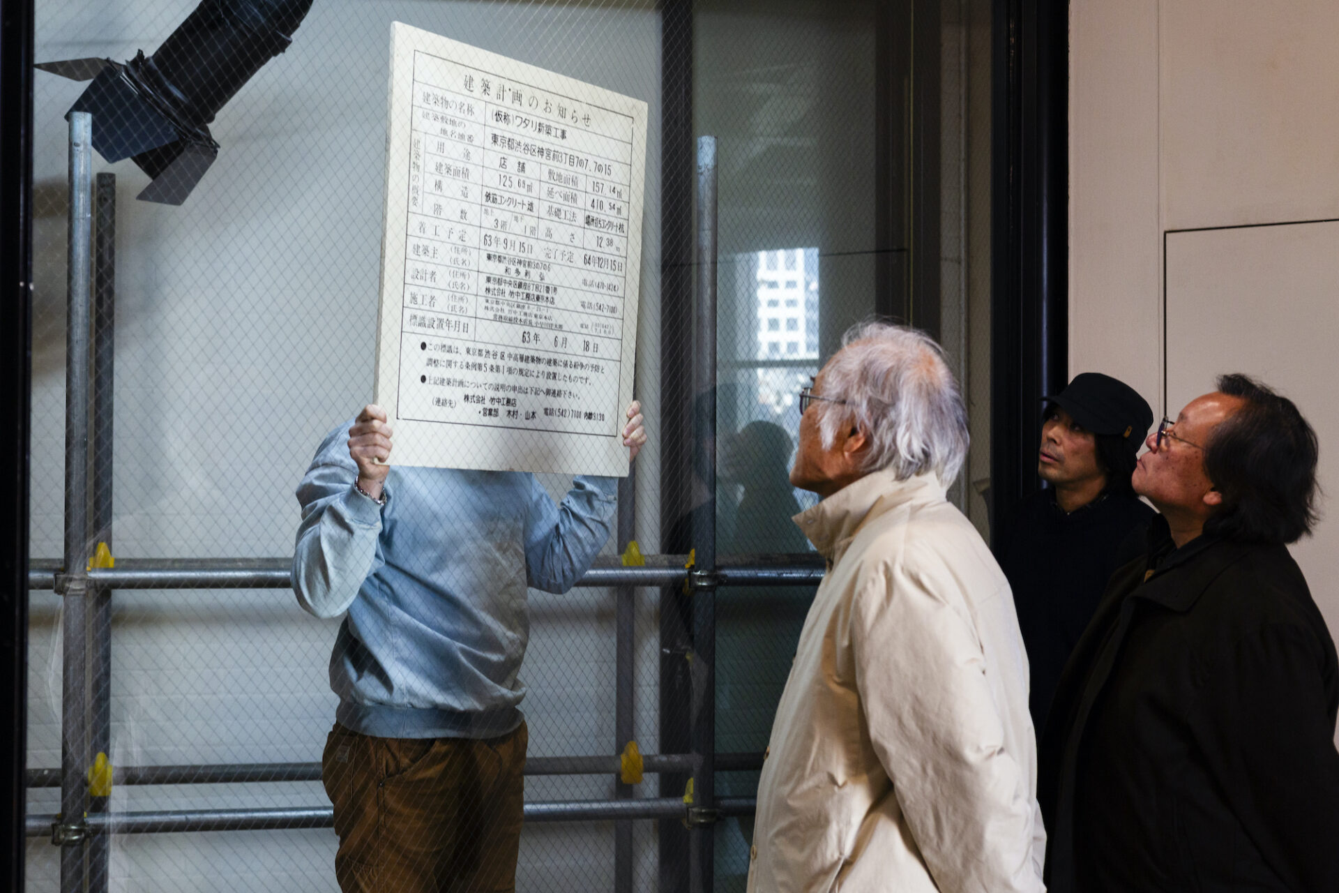 足場や金網フェンスの前で、3人の男性が「建築計画のお知らせ」と書かれた大判の紙を掲げ、内容を確認している。左の人物は顔が紙で隠れており、白髪の男性ともう一人の男性が紙を見上げている。