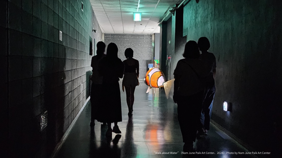 薄暗い廊下を、数人の人物がシルエットとなって奥へと歩いている。廊下の中ほどには光るクマノミ型の風船が浮かんでおり、オレンジ色の光を放っている。画面下部に「
