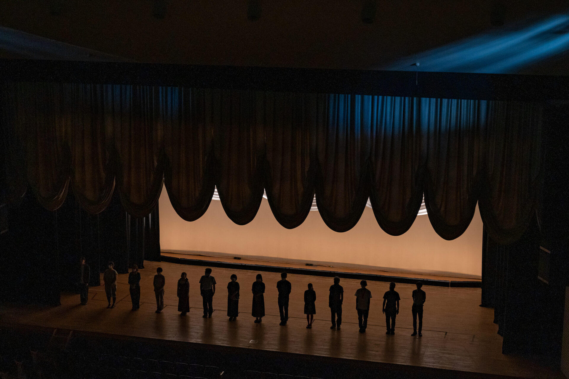 『9月0才』の写真。
薄暗い劇場の舞台上に、十数人の出演者がシルエットとなって横一列に立っている。背後には緩やかな弧を描く黒い緞帳が垂れ下がり、その隙間から温かみのある白い光が漏れている。上方から青みがかったスポットライトが斜めに差し込んでいる。