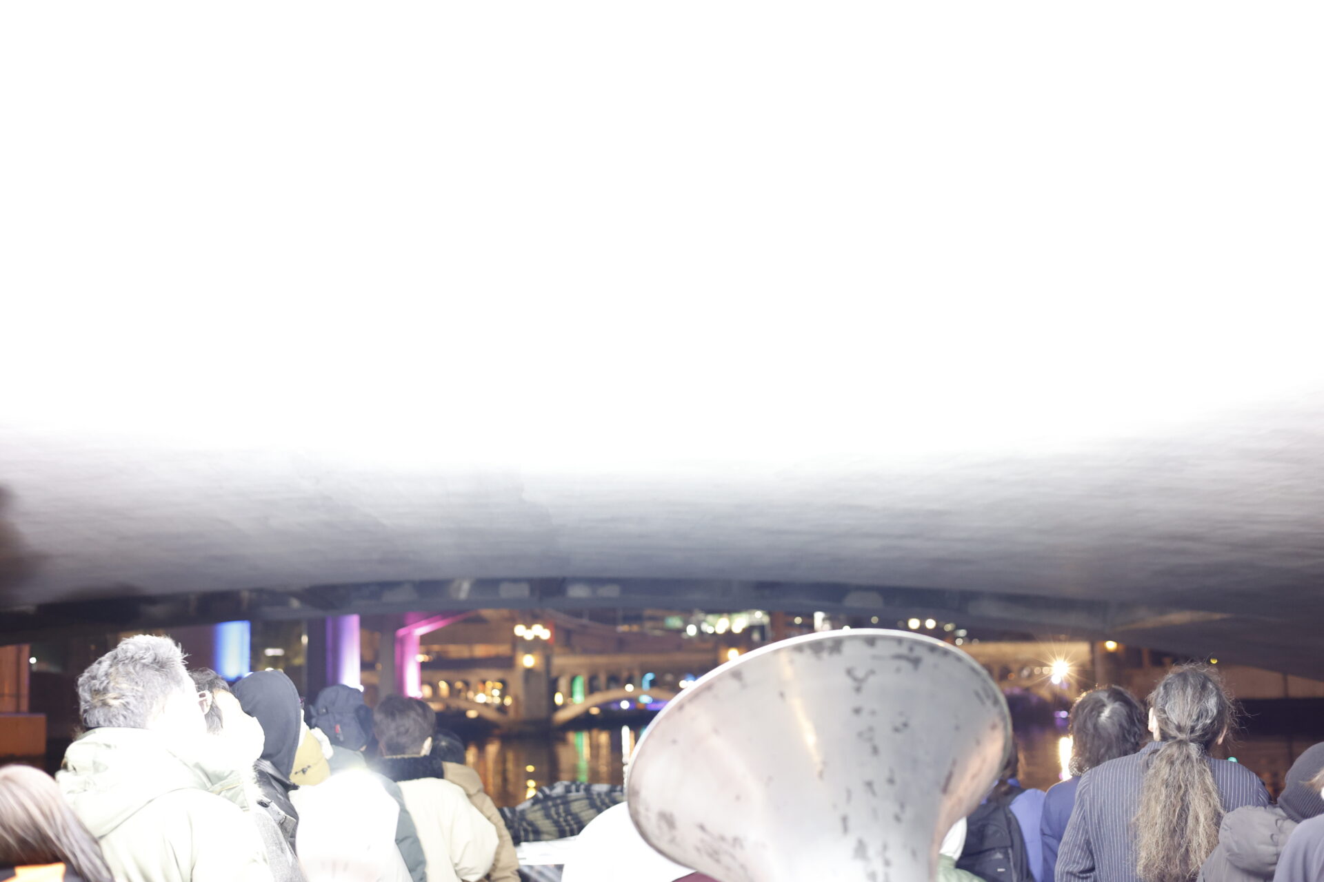 夜景の川岸か橋の下と思われる屋外で、多くの観客がシルエットとなって何かを見上げている。中央やや右にはくすんだ金属製の円錐形オブジェが見える。背景には橋やビルの夜景が広がり、青・紫・緑の照明が水面に反射している。上部は強い白い光で飛んでいる。