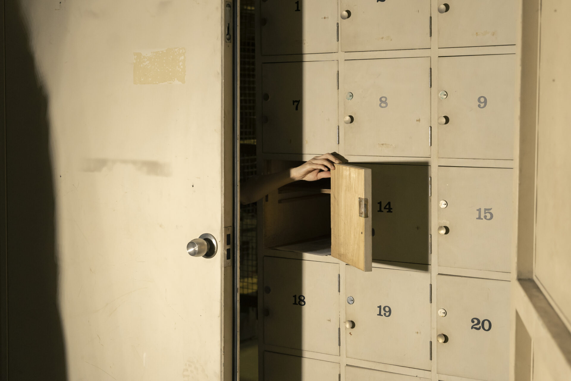 縦長の収納ロッカーが並ぶ薄暗い空間で、番号「14」のロッカー扉が開き、中から手が伸びて木製のボードを支えている。ロッカーには7〜20番台の番号が振られており、日差しが斜めに差し込んでいる。