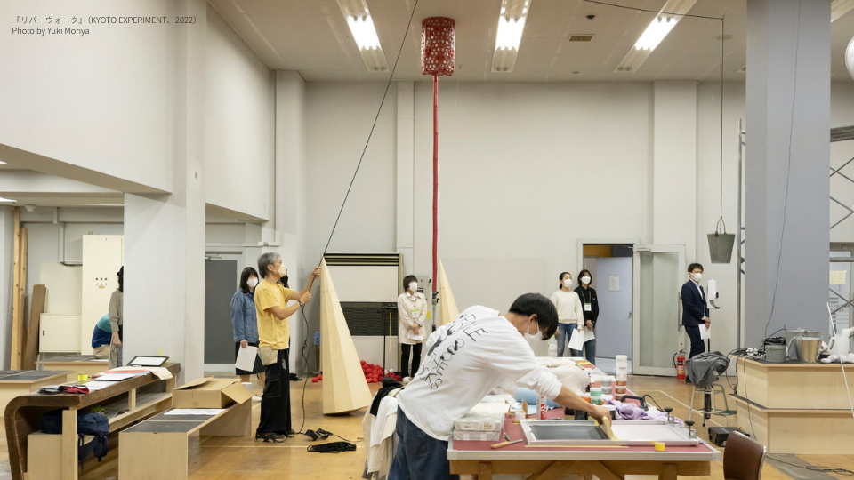 広い稽古場の室内で、天井から赤い提灯型のオブジェが吊られており、黄色いシャツの人物がその紐を引いている。手前ではもう一人が台上で作業中。奥には複数の観察者が立っており、三角形の造形物も見える。