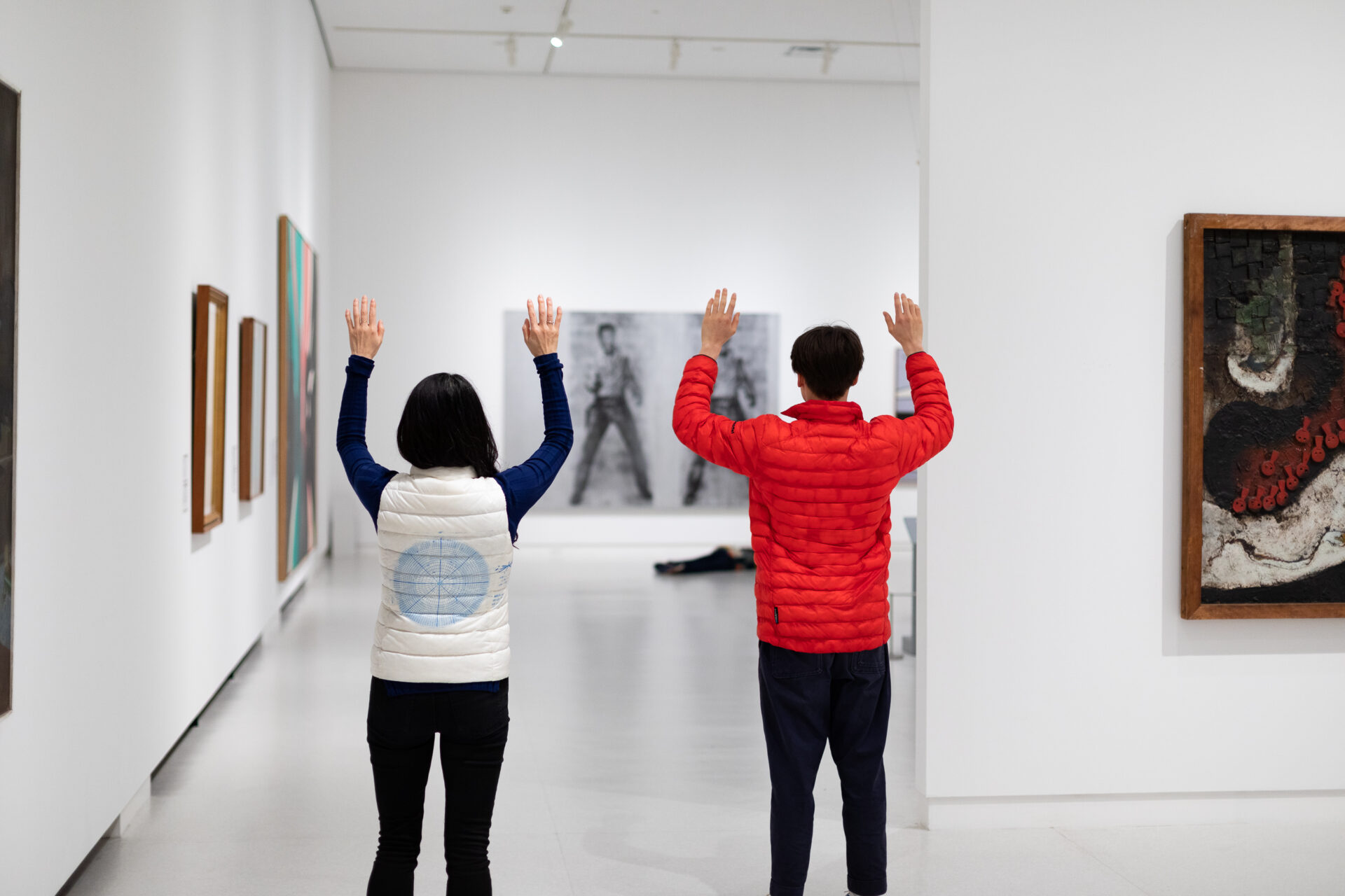 白い壁面に絵画が並ぶ美術館のギャラリー内で、白いベストを着た女性と赤いダウンジャケットを着た男性の二人が、背を向けて両手を高く掲げている。正面奥にはモノクロの大型写真作品が展示されている。
