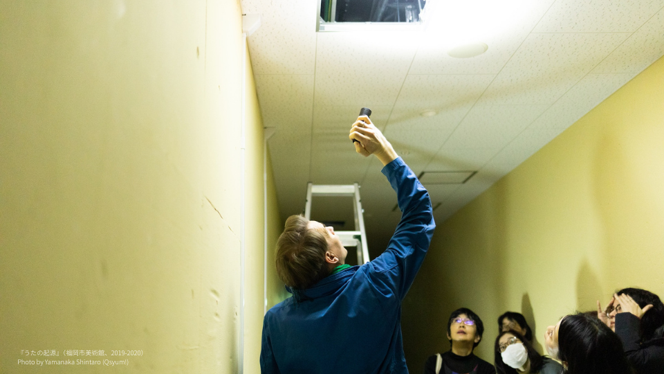 薄い黄色の壁が続く狭い廊下で、青いジャケットを着た人物が天井の点検口に向かって懐中電灯を照らし上げている。周囲には数人の観客が集まり、上を見上げている。奥には脚立が立てかけられている。左下にクレジット表記あり。