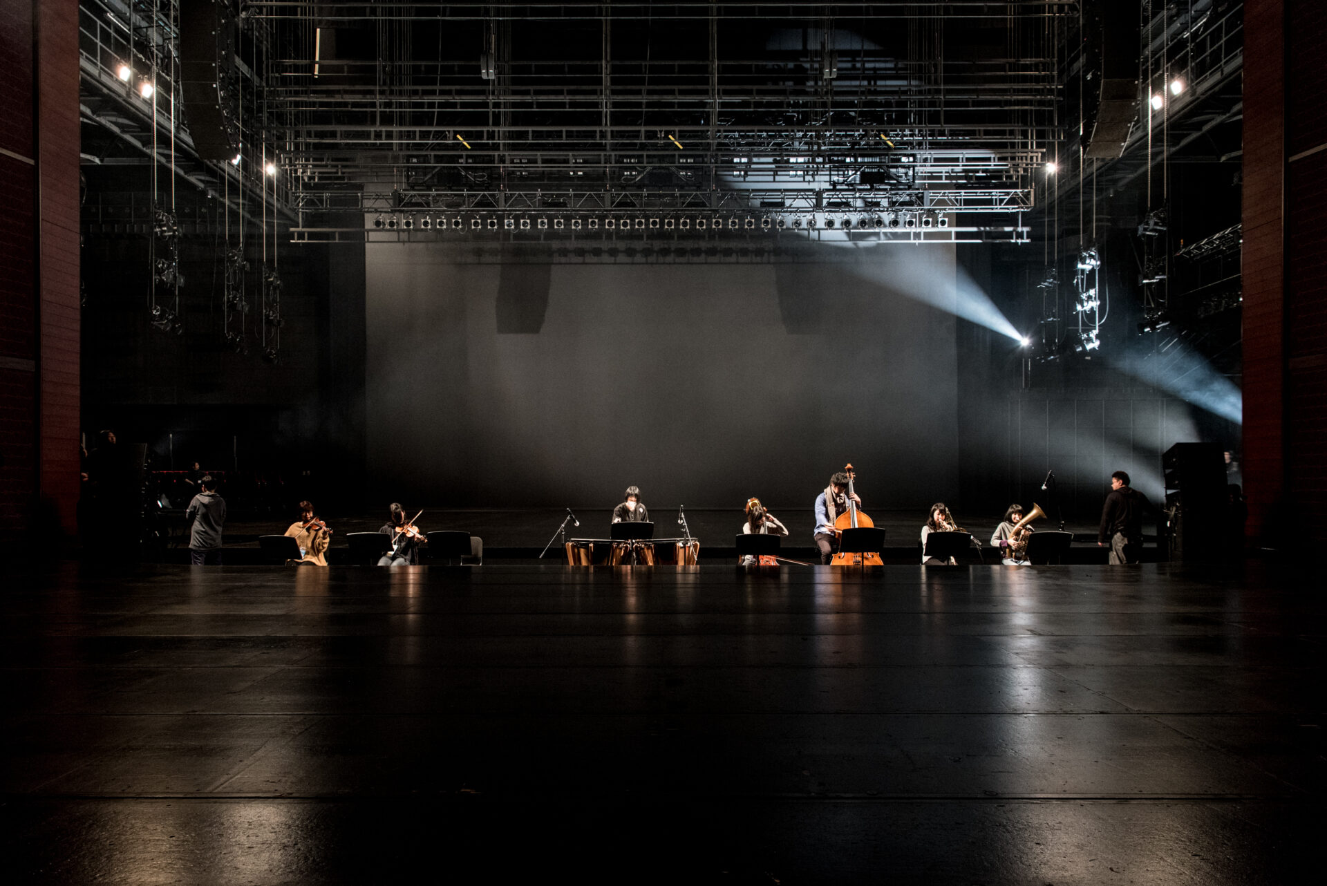 広大な暗い舞台上に、弦楽器・管楽器奏者ら約8〜9名が横一列に座り演奏している。舞台上方には無数のバトンや照明機材が吊られており、右側から強いスポットライトが斜めに差し込んでいる。