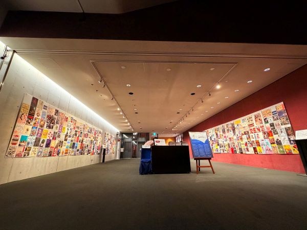 薄暗い照明の展示室。左右のコンクリート壁には、色とりどりの演劇・舞台公演のチラシが天井近くまでびっしりと貼り並べられている。室内中央やや左寄りに黒いテーブル状の受付カウンターがあり、その周辺にパンフレット類が置かれている。右手前には木製のイーゼルに縦長のパネルが立てかけられており、「座・高円寺×ギャラリーおちらしさん」の文字と展覧会情報が記載されている。