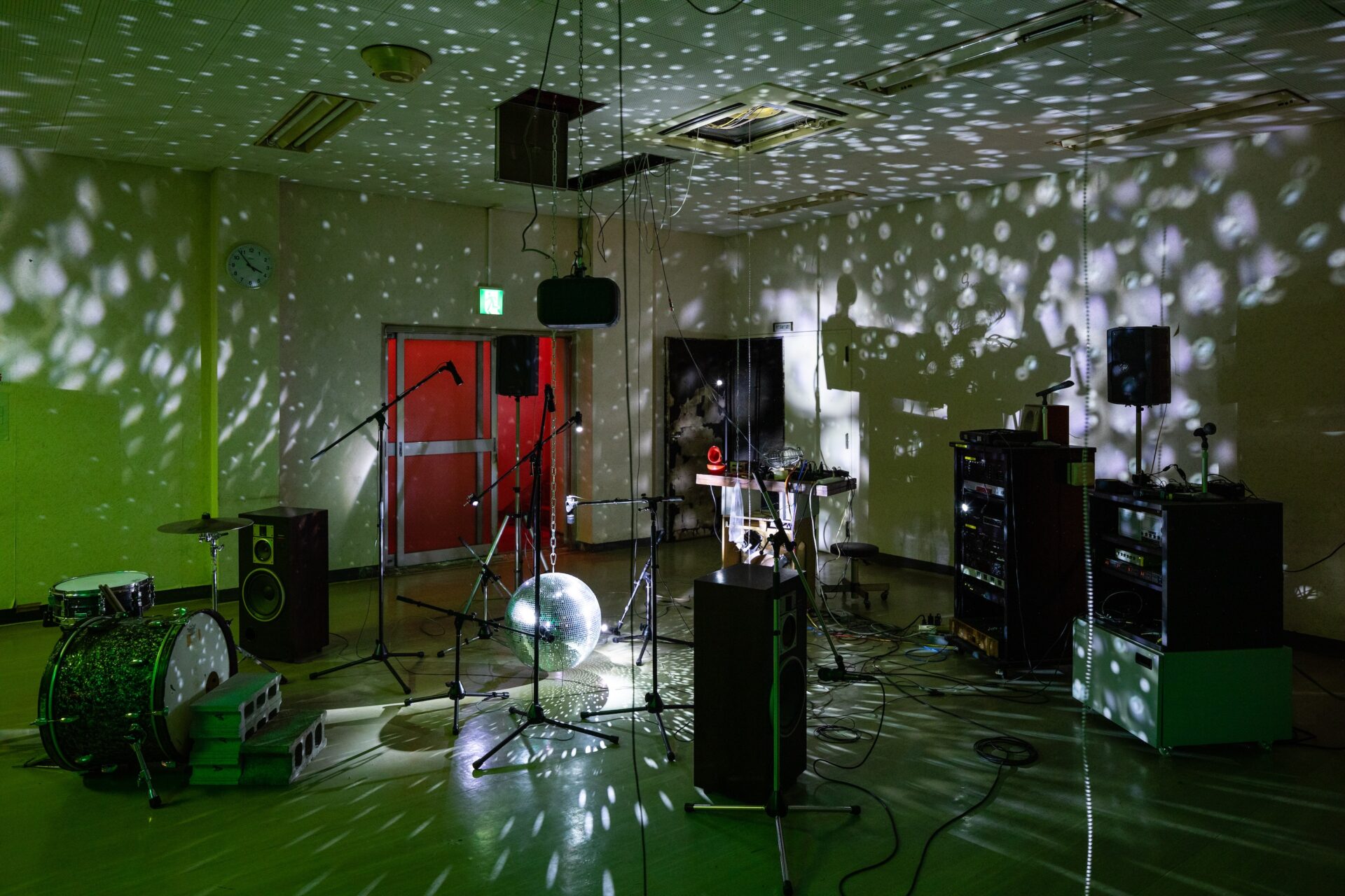 『O階』の写真。薄緑色の光に満たされた室内に、ドラムセット、マイクスタンド、スピーカー、シンセサイザーなどの楽器・音響機材が散在している。中央には大きなミラーボールが床に置かれ、壁と天井には光の反射による白い斑点模様が広がっている。