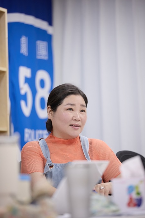 稽古場にて、劇団58ROUTEのロゴが描かれた青い看板を背景に、テーブルに向かって話すコ・スヒ。オレンジのニットにデニムのサロペットを着用。