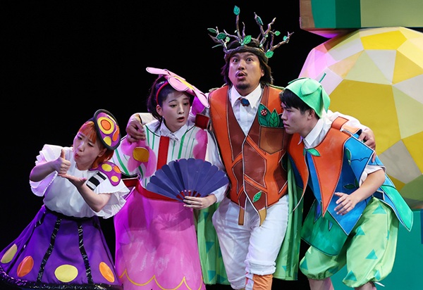 『夏の夜の夢』2025年公演の舞台写真。カラフルな衣装をまとった4人の出演者が身を寄せ合い、豊かな表情で演じている。中央の男性は木の枝をあしらった冠と茶色のベストを着用し、扇子を持つ女性や蝶・植物を模した衣装の俳優たちと並ぶ。